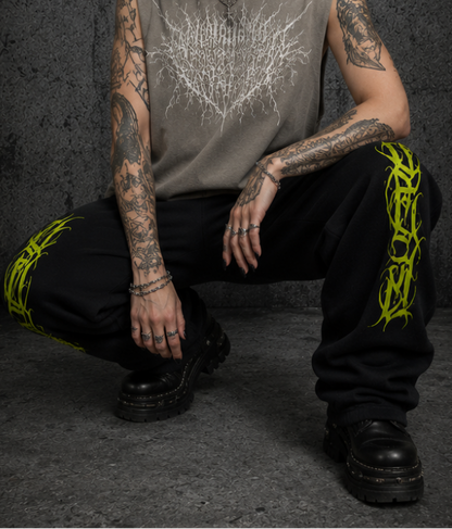 A model is shown squatting down wearing a sleeveless taupe-gray top with a white tree graphic, black pants with neon green and yellow calligraphy graphics,spelling venom culture, wearing black boots, posing against a dark textured concrete wall.