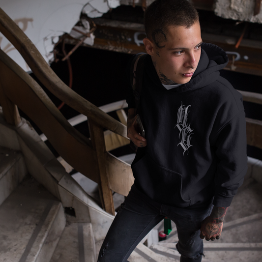 alternative male model with face tattoos stands on spiral staircase and shows fron view of venom culture hoodie featuring gothic calligraphy v.c logo
